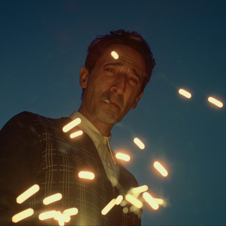 Adrien Brody smokes as sparks dance around him in a still from the movie The Brutalist