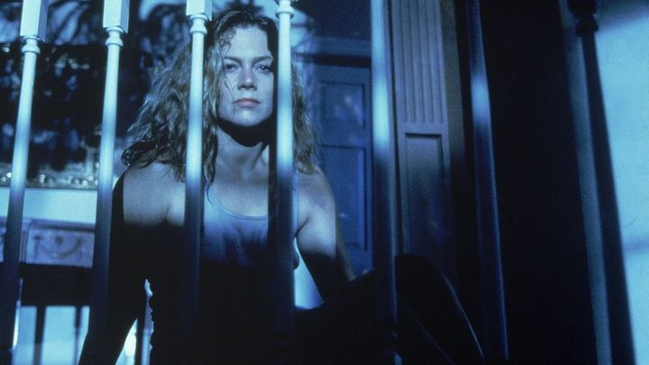 Kathleen Turner as Barbara Rose looking serious behind a bannister in a dark house in The War of the Roses.