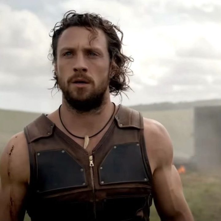 Aaron Taylor-Johnson looks sternly forward as a small fire rages behind him in a still from the movie Kraven the Hunter.