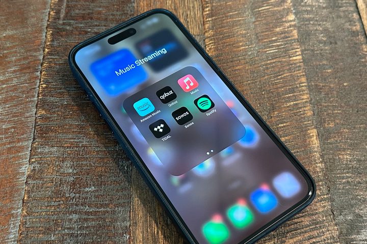 An iPhone 15 Pro Max on a wooden table displaying streaming apps Amazon Music, Qobuz, Apple Music, Tidal, Sonos, and Spotify.