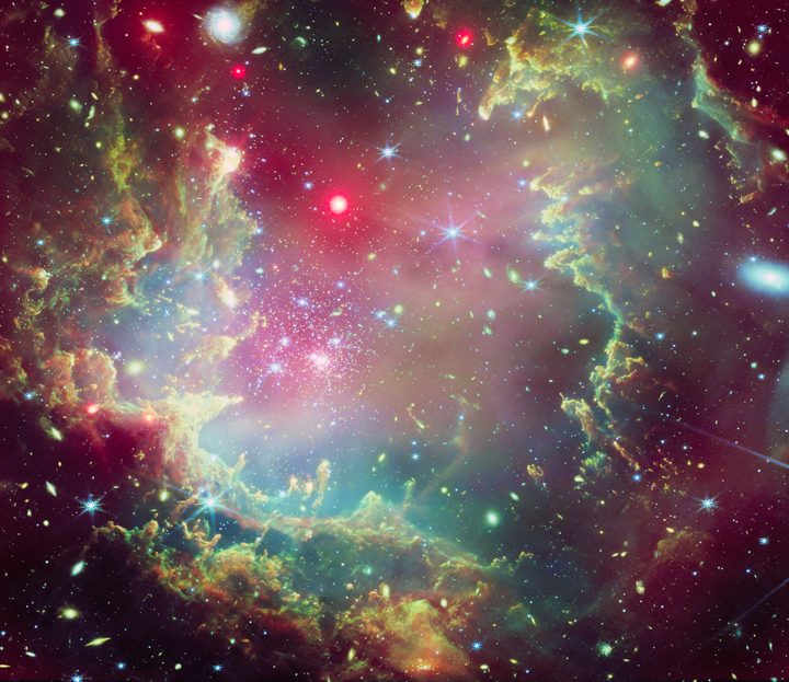 This image depicts star cluster NGC 602 in vibrant and festive colors. The cluster includes a giant dust cloud ring, shown in greens, yellows, blues, and oranges. The green hues and feathery edges of the ring cloud create the appearance of a wreath made of evergreen boughs. Hints of red representing X-rays provide shading, highlighting layers within the wreath-like ring cloud.