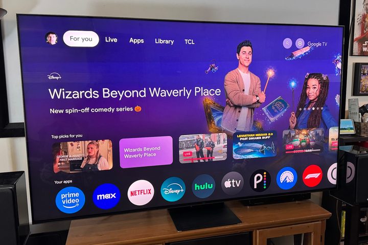 The 75-inch 2024 TCL QM8 mini-LED TV on a wooden home theater credenza displaying the Google TV home screen.