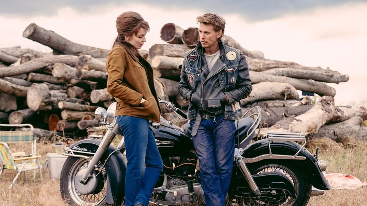 A woman and man talking while leaning against a motorcycle in The Bikeriders.