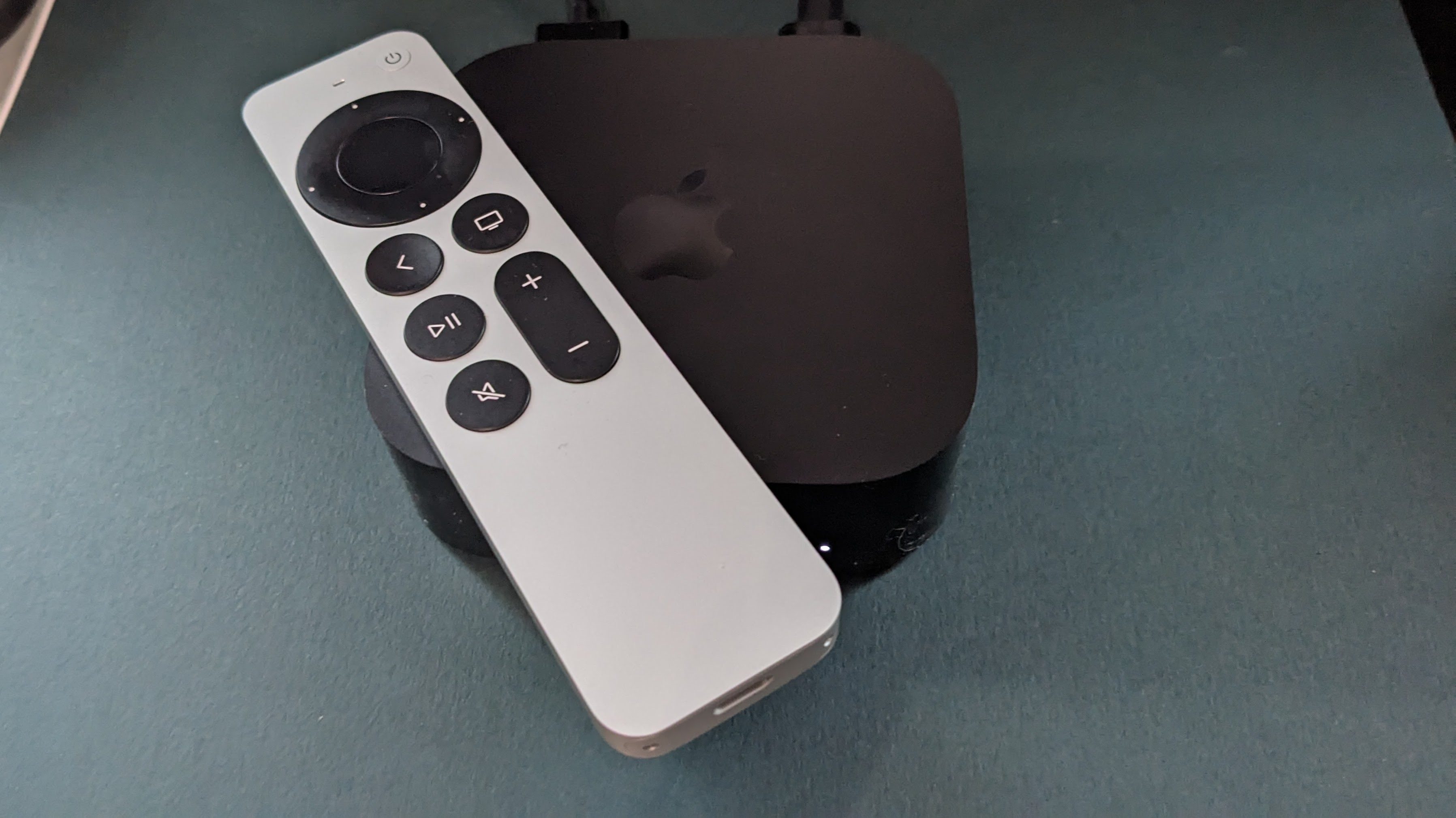 An Apple TV 4K sits against a green background. The silver apple remote sits on top of the streaming box.