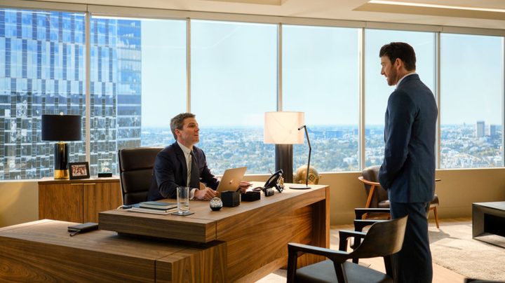 A man sits at his desk as another one stands in front of it in Suits LA.