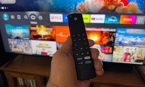 A hand holding the Amazon Fire TV remote in front of the Amazon Fire TV Omni Mini-LED TV.