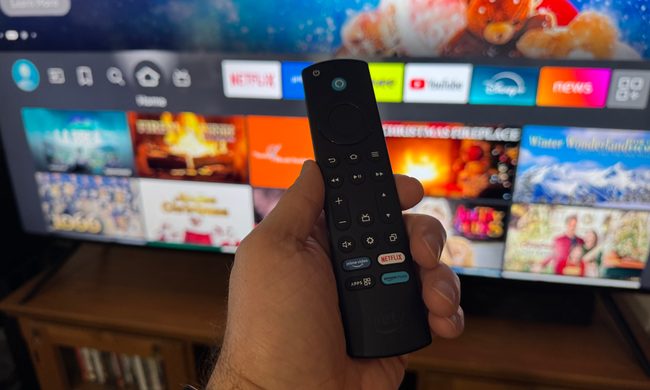 A hand holding the Amazon Fire TV remote in front of the Amazon Fire TV Omni Mini-LED TV.