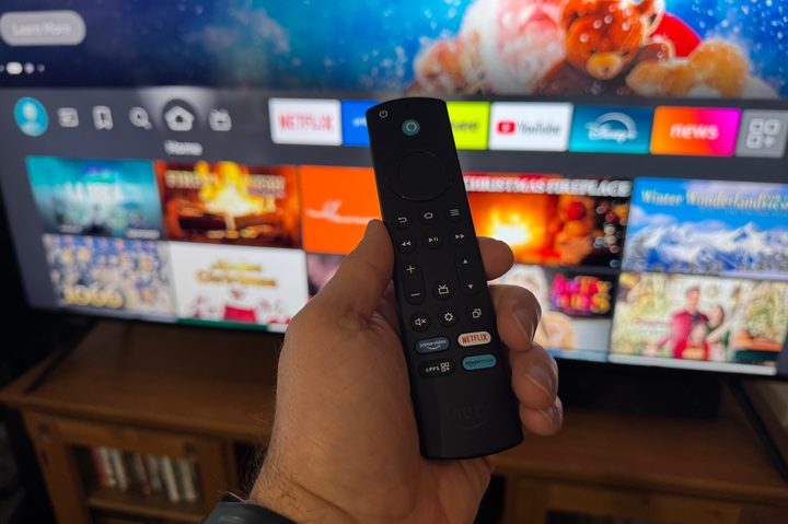 A hand holding the Amazon Fire TV remote in front of the Amazon Fire TV Omni Mini-LED TV.