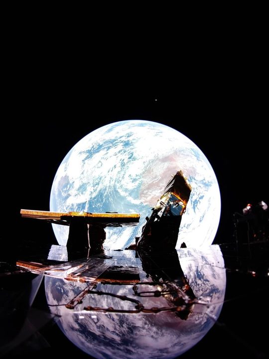 Firefly’s Blue Ghost lander captures image of Earth reflecting off the solar panel with the Moon on the horizon above Earth. Firefly’s X-band antenna and NASA’s LEXI payload are also shown on the top deck of the lander.