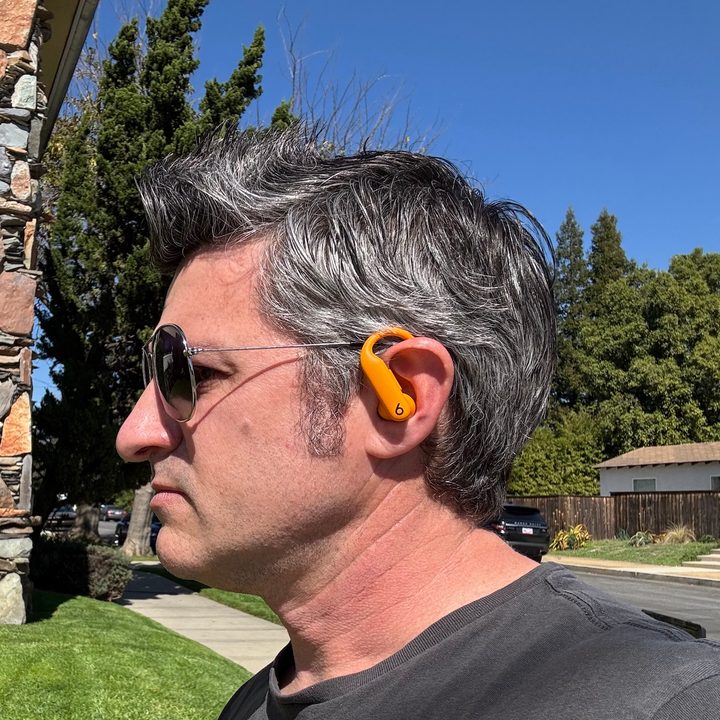 A man wearing the Beats Powerbeats Pro 2 earbuds in Electric Orange outside on a sidewalk.