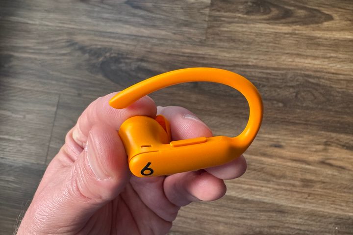 A hand holding an orange Beats Powerbeats Pro 2 earbud, showing the physical buttons.