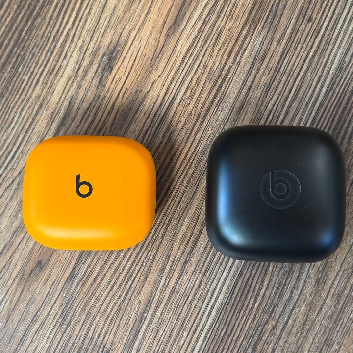 A Beats Powerbeats Pro 2 case in orange next to a Powerbeats Pro case in black on a wooden floor photographed from above.
