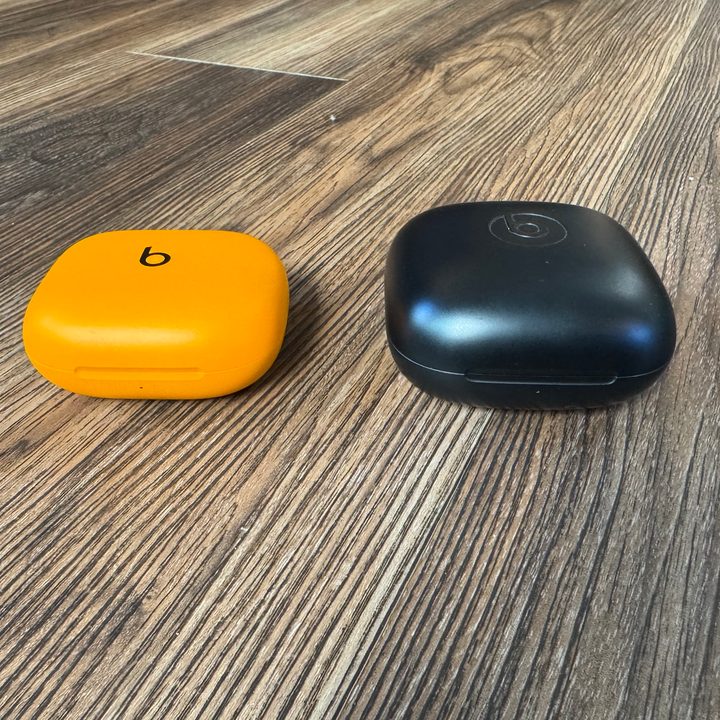 A Beats Powerbeats Pro 2 case in orange next to a Powerbeats Pro case in black on a wooden floor photographed from a low-profile angle.