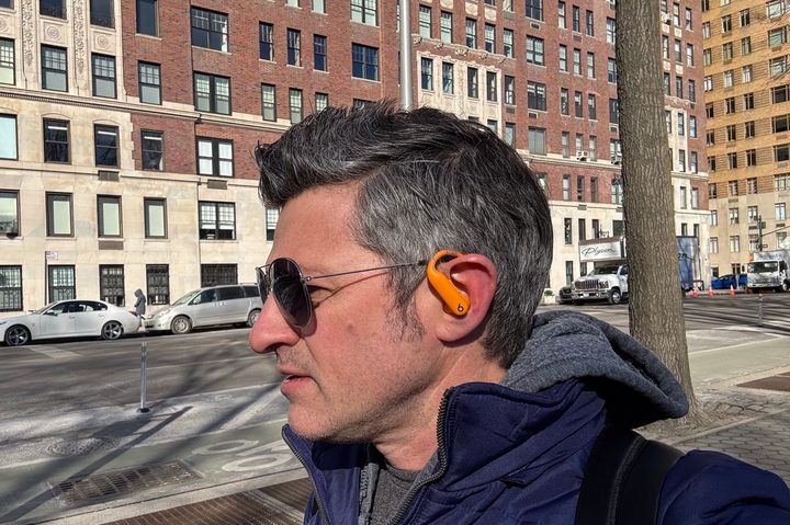 A man in a blue jacket and sunglasses wearing orange Beats Powerbeats Pro 2 earbuds while walking on a New York City street.