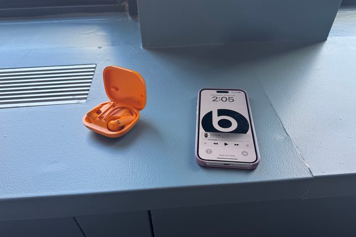 A orange pair of Beats Powerbeats Pro 2 headphones in their case next to an iPhone displaying the Beats logo.