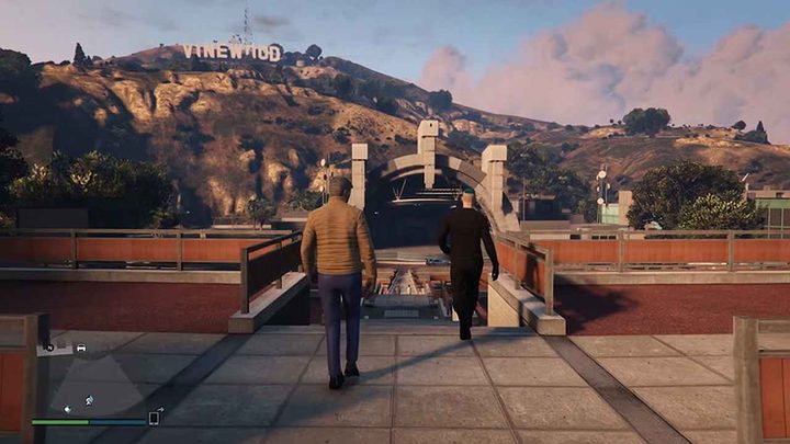 A figure wearing a tan jacket and jeans walks alongside a figure with blue hair wearing all black. They walk down towards a large open ampitheater. A sign that reads "Vinewood" stands behind the venue.
