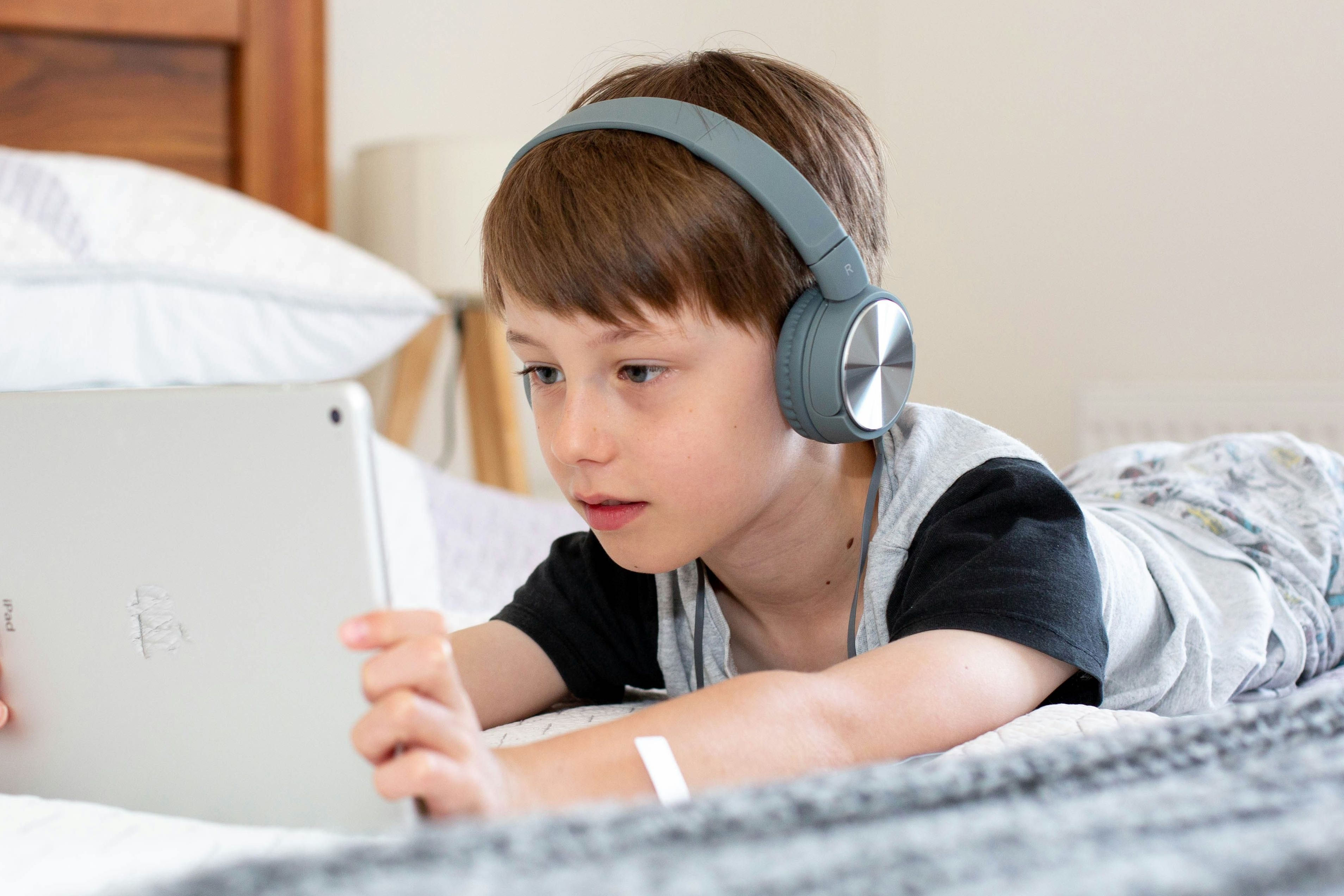 Kid lying on a bed wearing headphones while using a tablet.