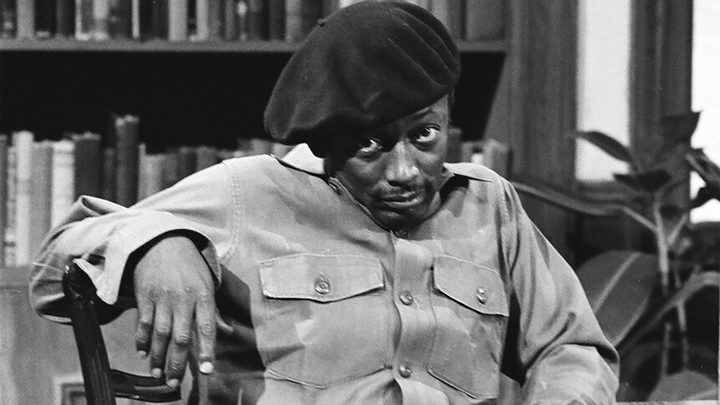 A black and white image of Garrett Morris from SNL sitting in a chair with a hat, head down looking up.