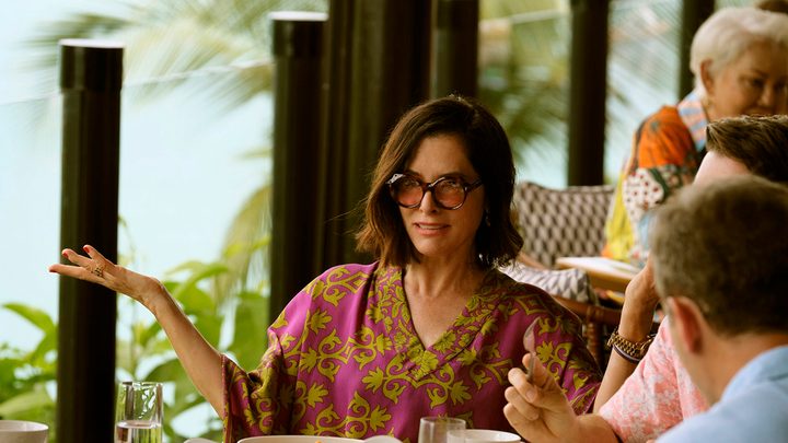 A woman sits at a resort with a floral dress and sunglasses gesticulating with her arms in The White Lotus season 3.