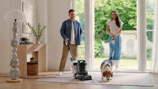 man and woman vacuuming rug while dog runs through room