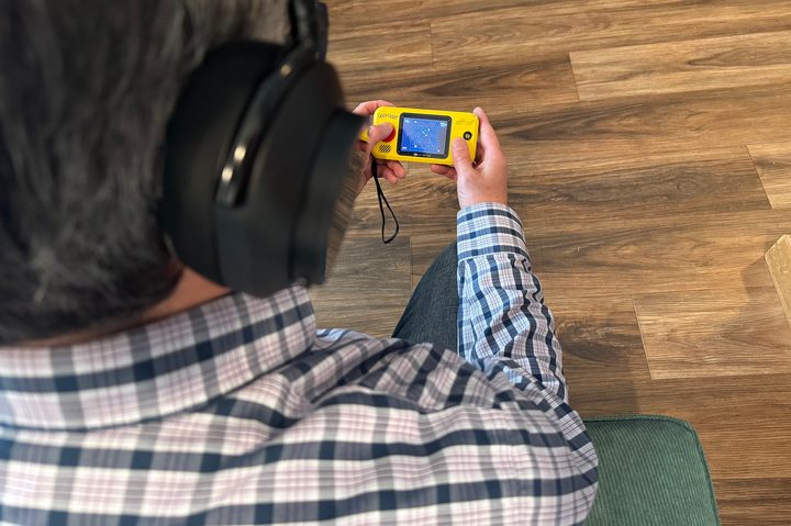 An over-shoulder angle of a man sitting on a couch wearing teh Master & Dynamic MW75 Neuro headphones while playing a handheld game.