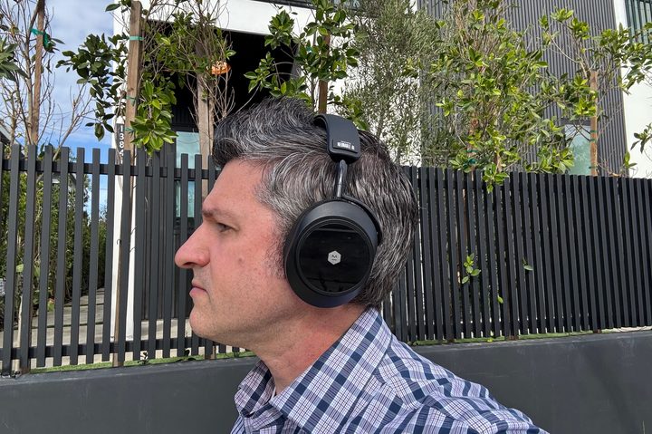A man wearing the Master & Dynamic MW75 Neuro headphones outside with a black fence behind him.