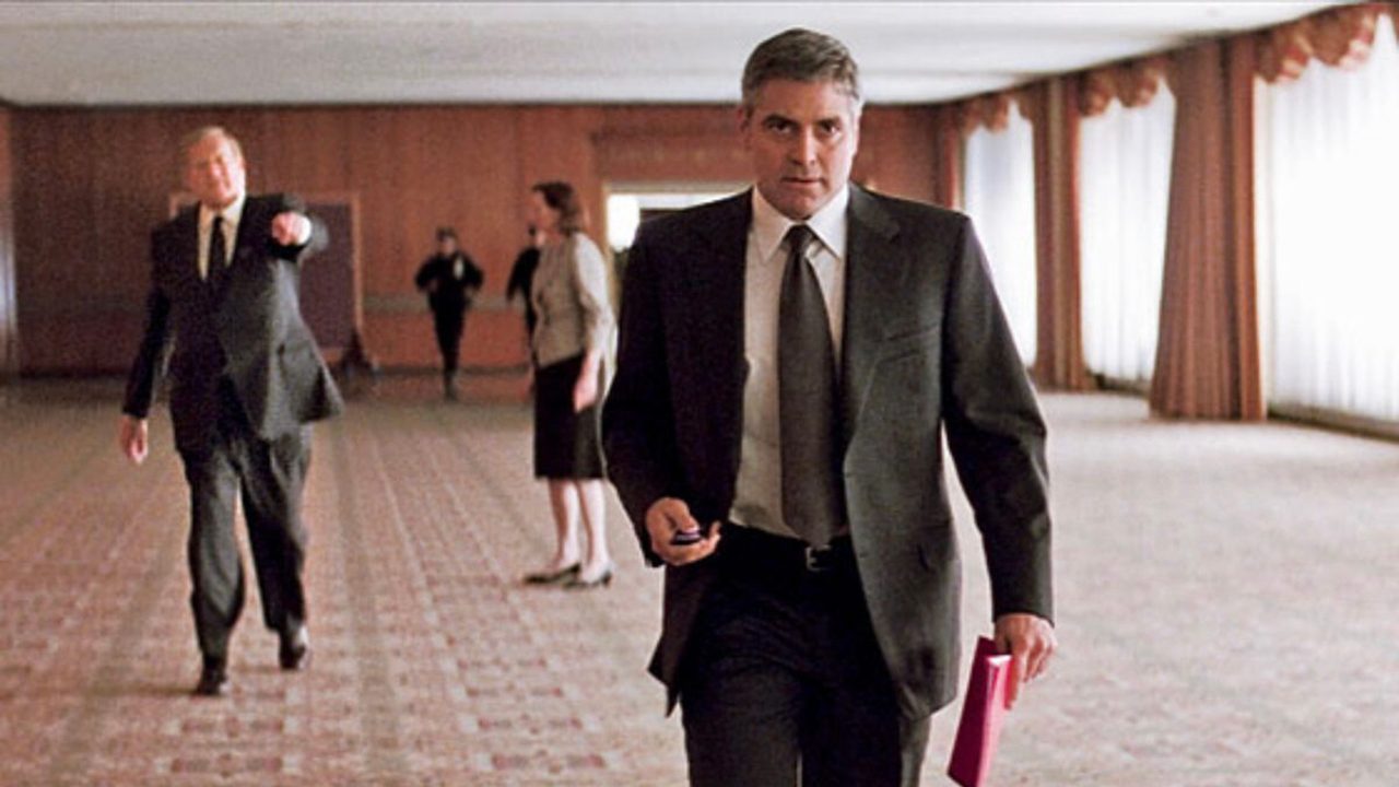 George Clooney in a suit walking down a hallway as a man walks after him pointing in Michael Clayton.