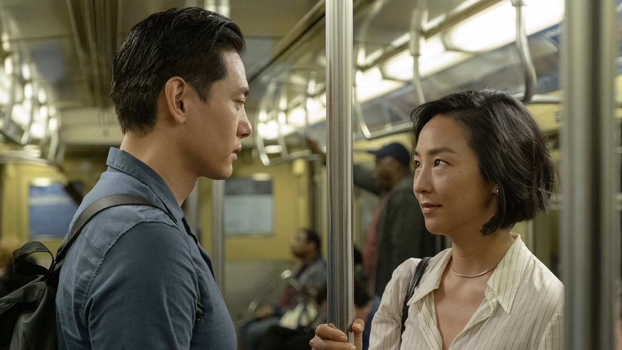 A woman and man look lovingly at one another while standing on a subway car in Past Lives.