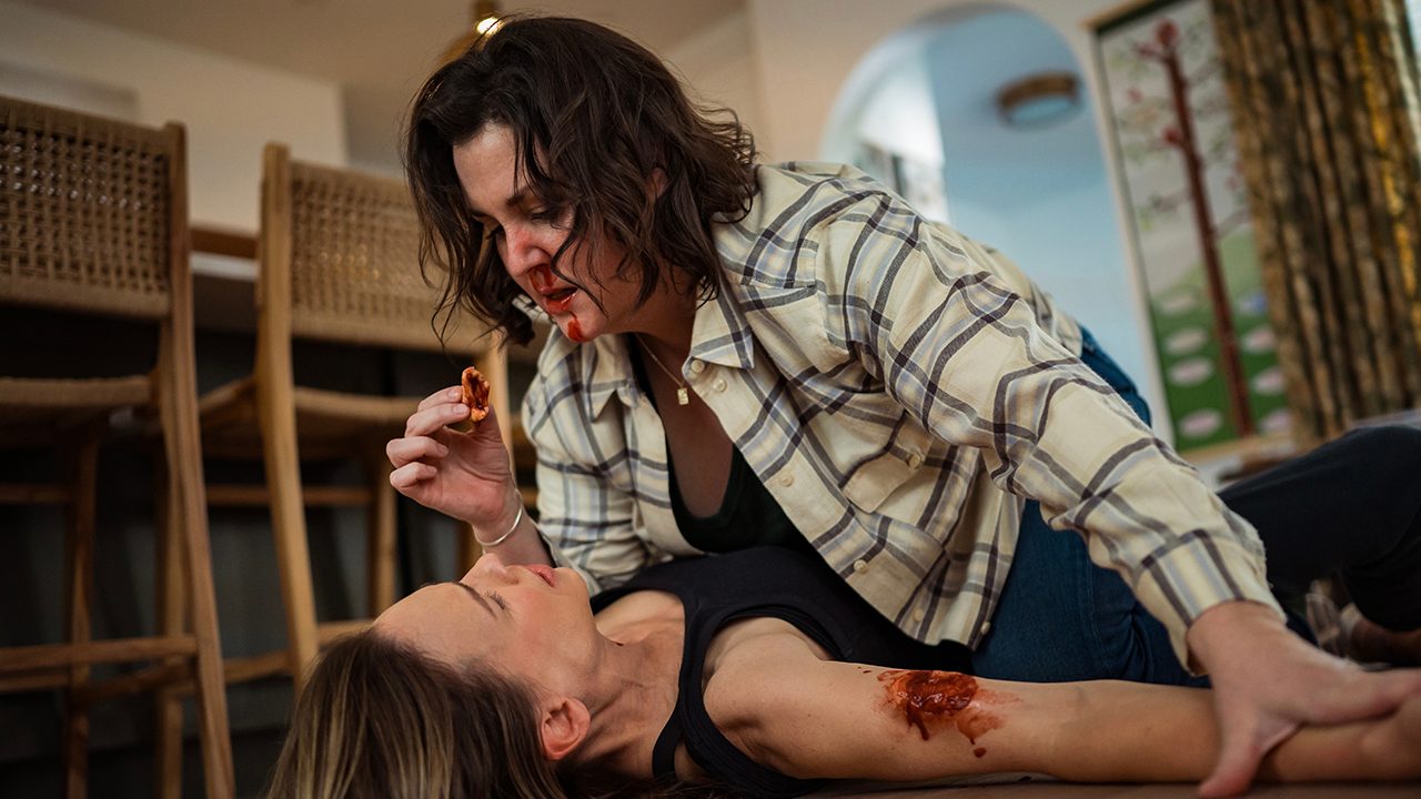 Shauna pinning Melissa down to the ground, holding a piece of skin from her arm over her mouth, Shauna's mouth bloody in Yellowjackets.