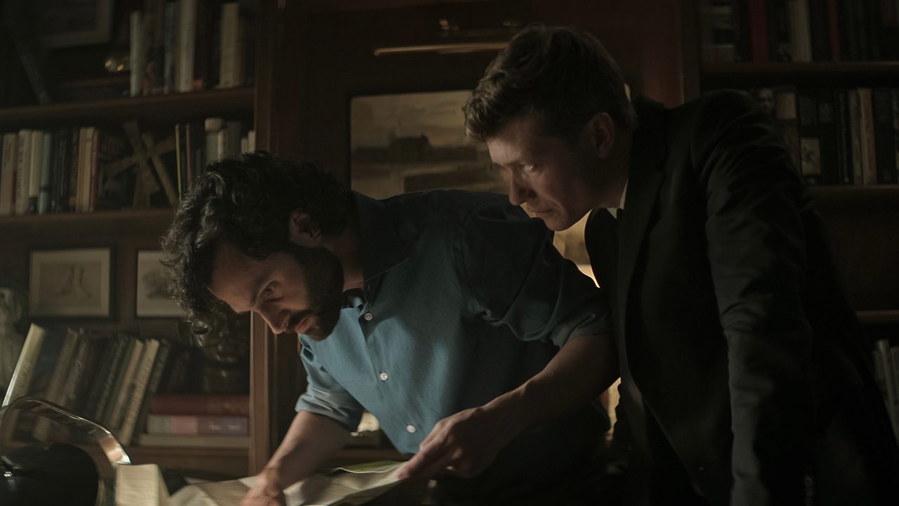 Joe looking at something on a table as Rhys hovers behind him in You.