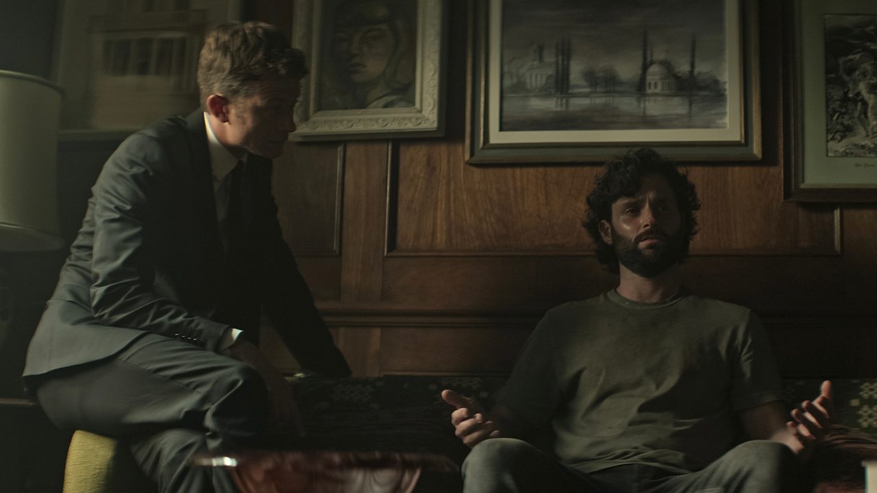 Joe sitting on the couch, hands up looking terrified as Rhys looks on in You.
