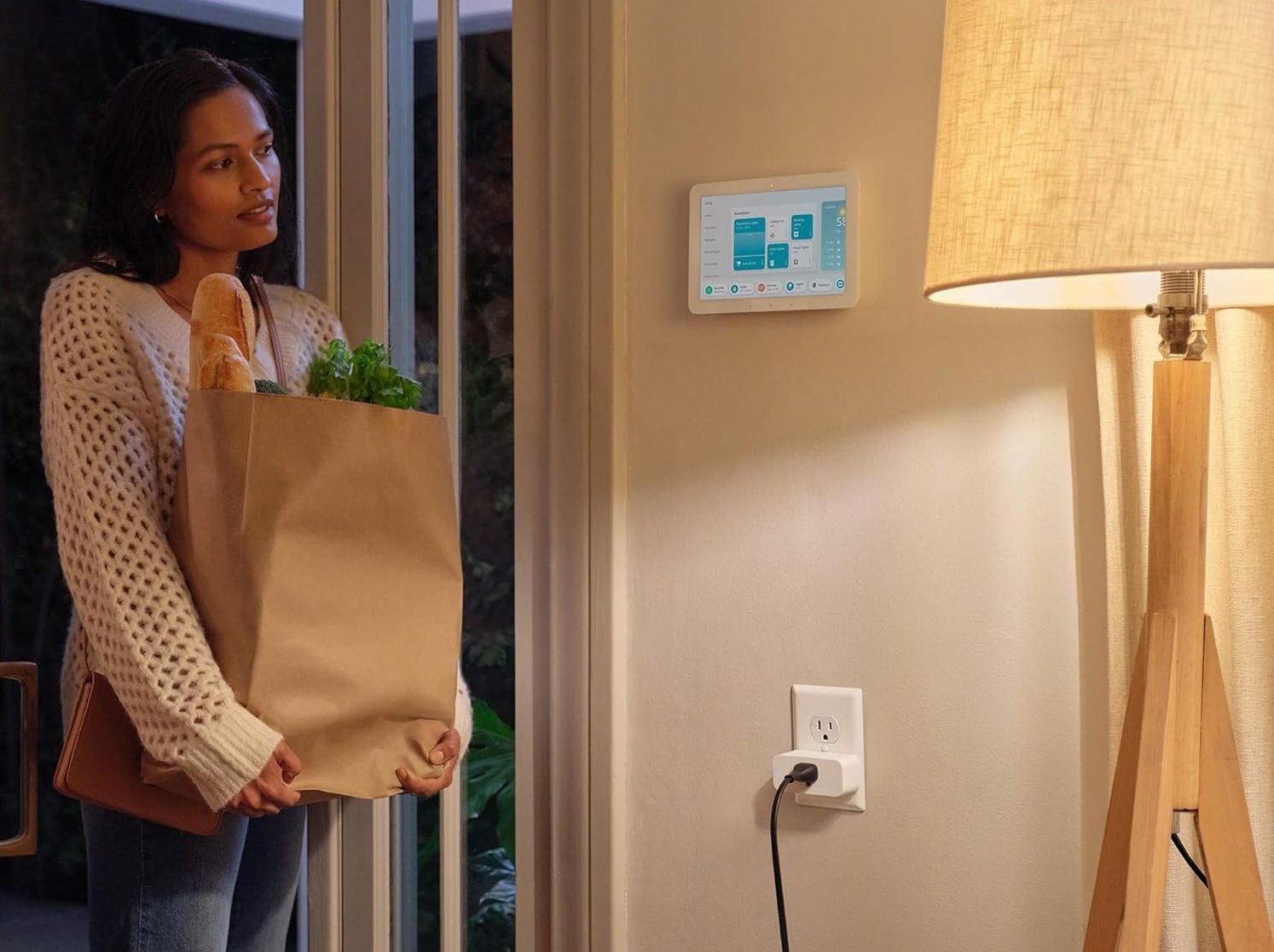 A woman standing in her home carrying groceries and looking at her Amazon Smart Plug.