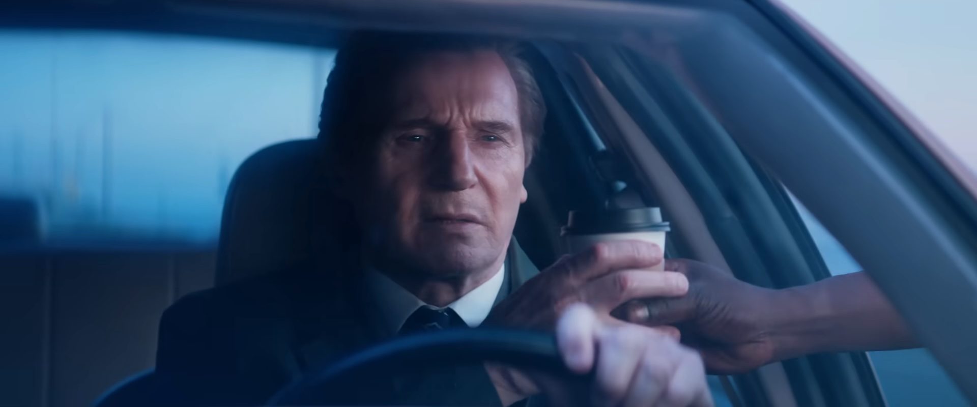 Liam Neeson as Frank Drebin Jr. holding a coffee cup in a car in The Naked Gun.