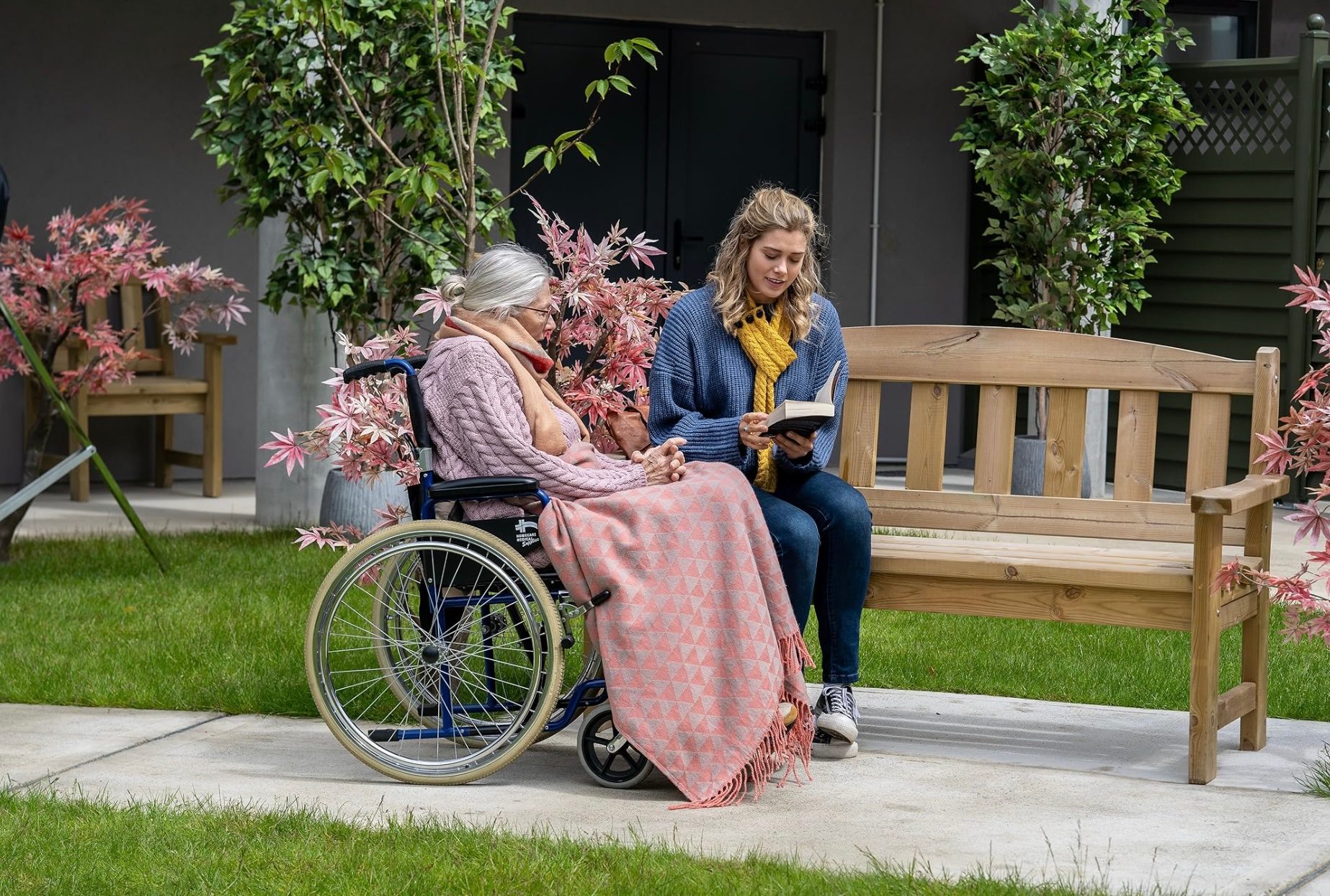A girl reads to a woman in a wheelchair.