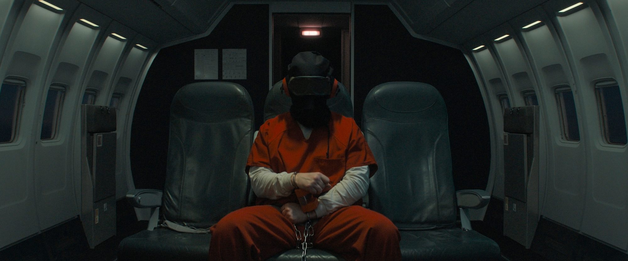 A blindfolded prisoner sits on a plane.