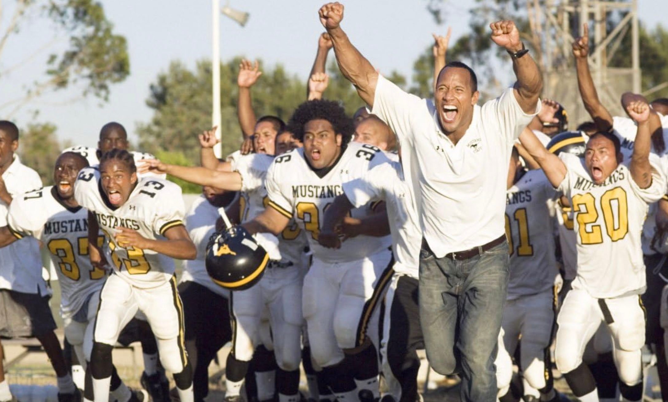 Dwayne Johnson and football players throw their hands up.