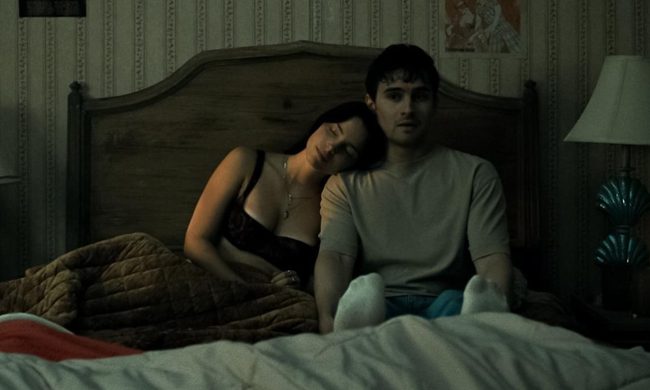 Nikki (Inde Navarette) and Bear (Michael Johnston) sitting on a bed together in the horror film, Obsession, written and directed by Curry Barker.