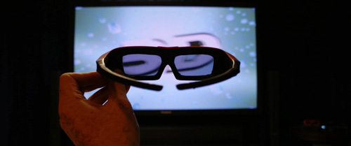 A hand holding a set of 3D glasses in front of a TV.