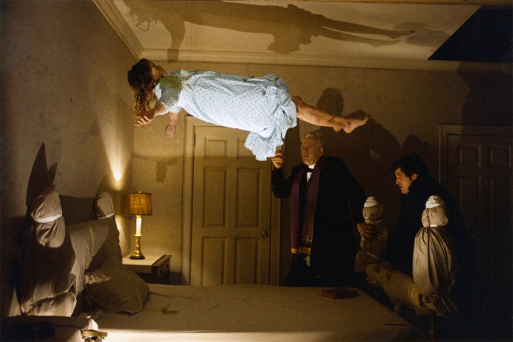 A girl levitates above her bed in front of two priests.