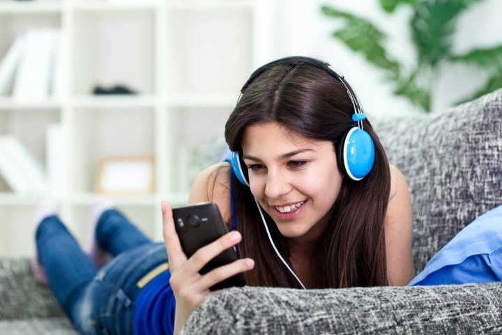 A woman wearing headphones looks at a smartphone while listening to music on the sofa.