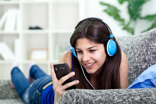 A woman wearing headphones looks at a smartphone while listening to music on a sofa.