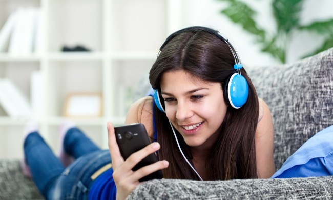 A woman wearing headphones looks at a smartphone while listening to music on a sofa.
