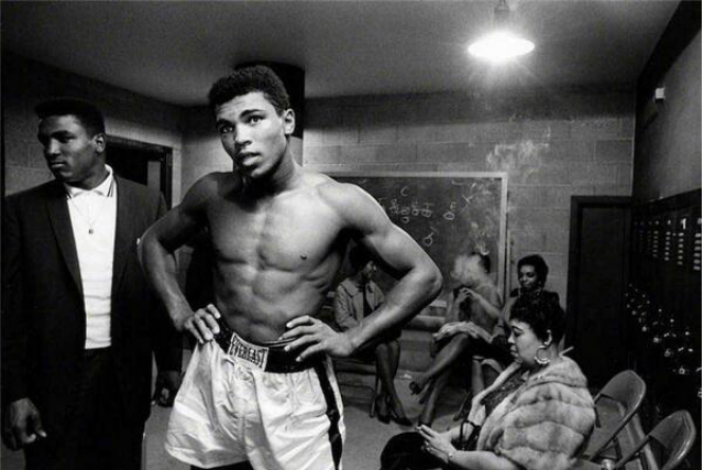Muhammad Ali está de pé com as mãos nos quadris.
