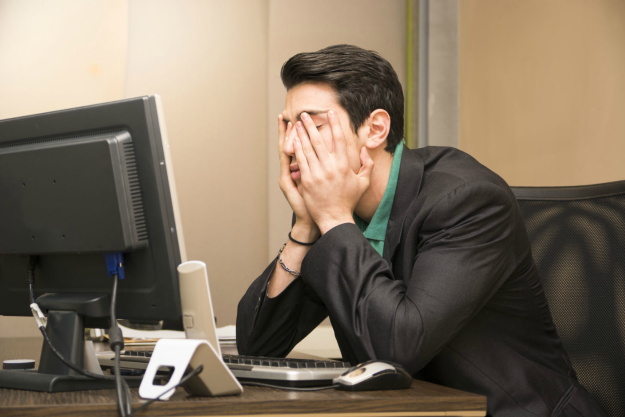 A person sitting at a desk, in front of a computer monitor with their head in their hands.
