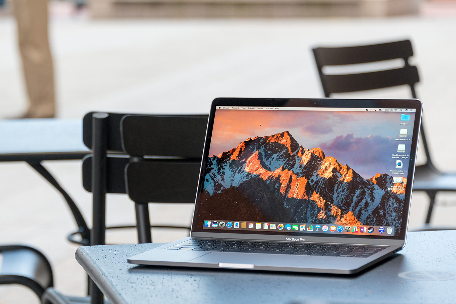 A MacBook Pro sitting on a desk with chairs behind it. The macOS Sierra operating system is on its screen.