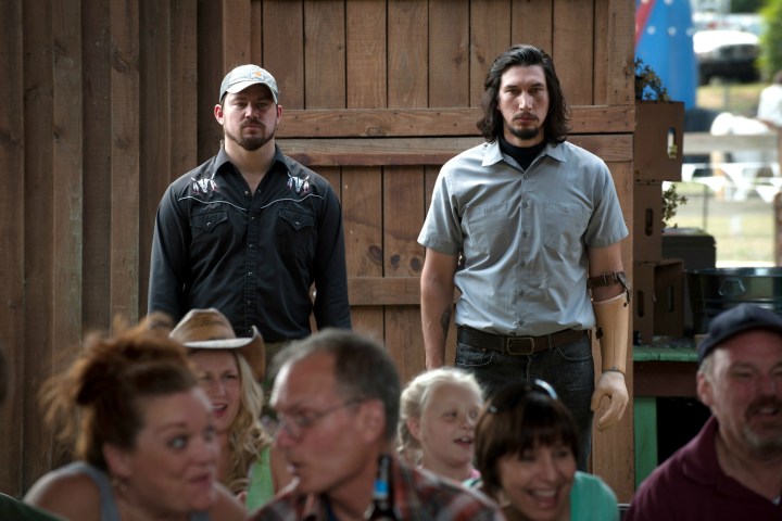 Channing Tatum and Adam Driver stand next to each other in the back of a room in Logan Lucky.