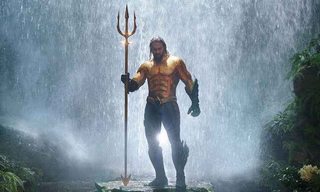 Aquaman standing in front of a waterfall holding a trident in the movie Aquaman.