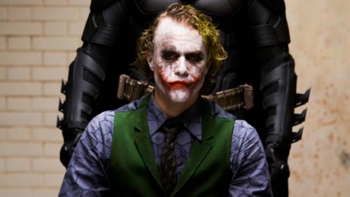 Heath Ledger como o Coringa sentado com Batman atrás dele em O Cavaleiro das Trevas.