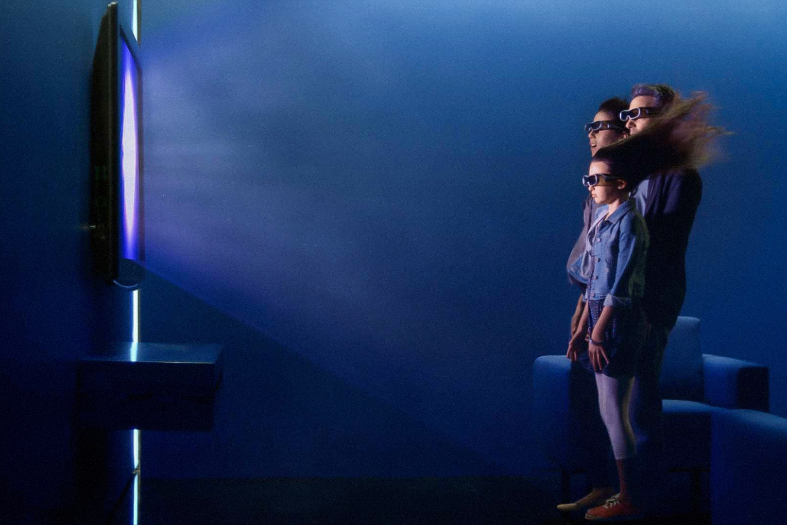 Three individuals stand in front of a large 3D TV wearing 3D glasses and having their hair blow back by the realism of the image. 
