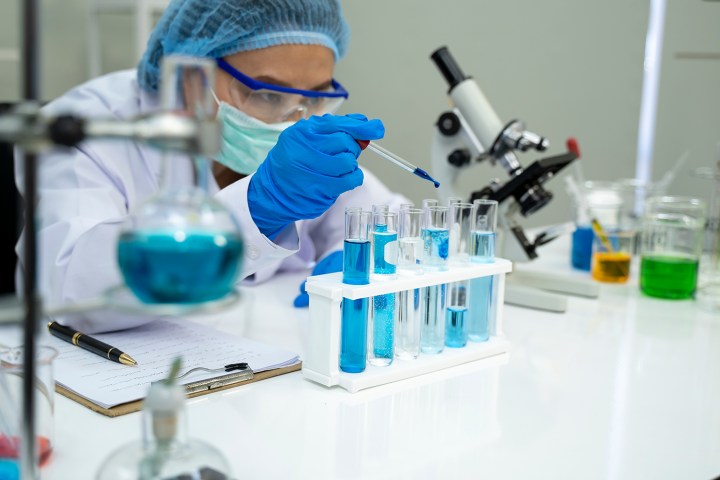 Científico en laboratorio trabajando con un cuentagotas, tubos de ensayo y líquido azul y rodeado de varios equipos de laboratorio.